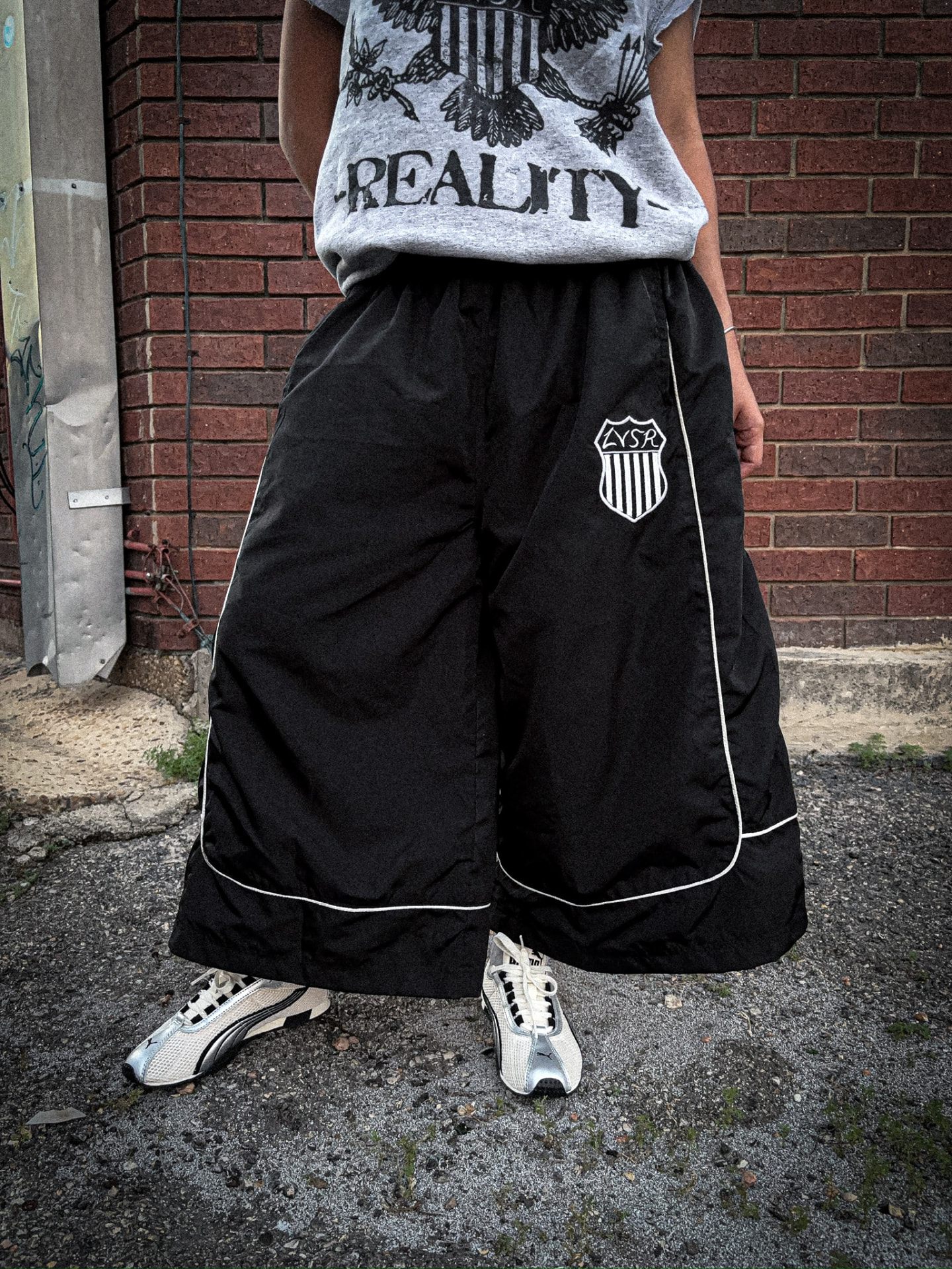 Model in grey LVSR Reality tee with black wide-leg shield pants, low angle view