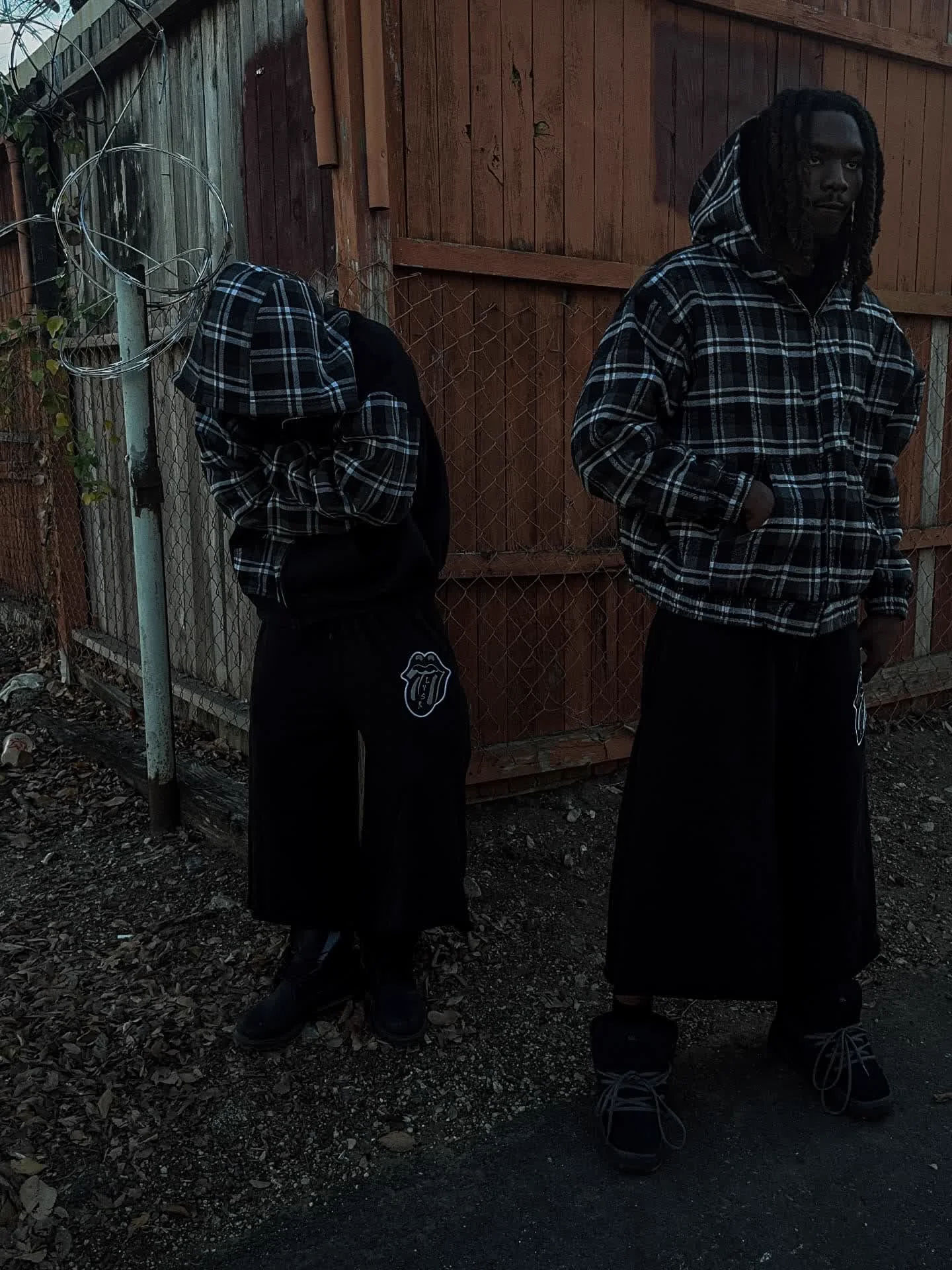 Two models in matching LVSR plaid hoodies and black wide-leg pants against a wooden fence